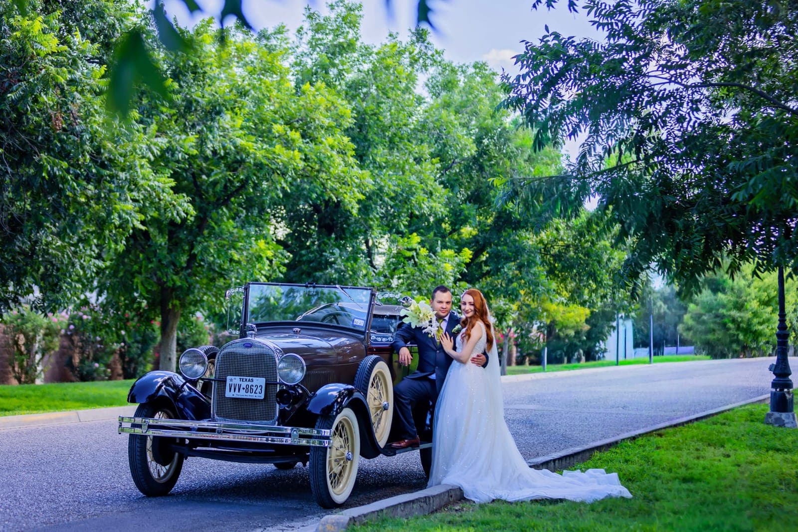 Pareja de novios posando junto a un coche clásico marrón en una calle arbolada.