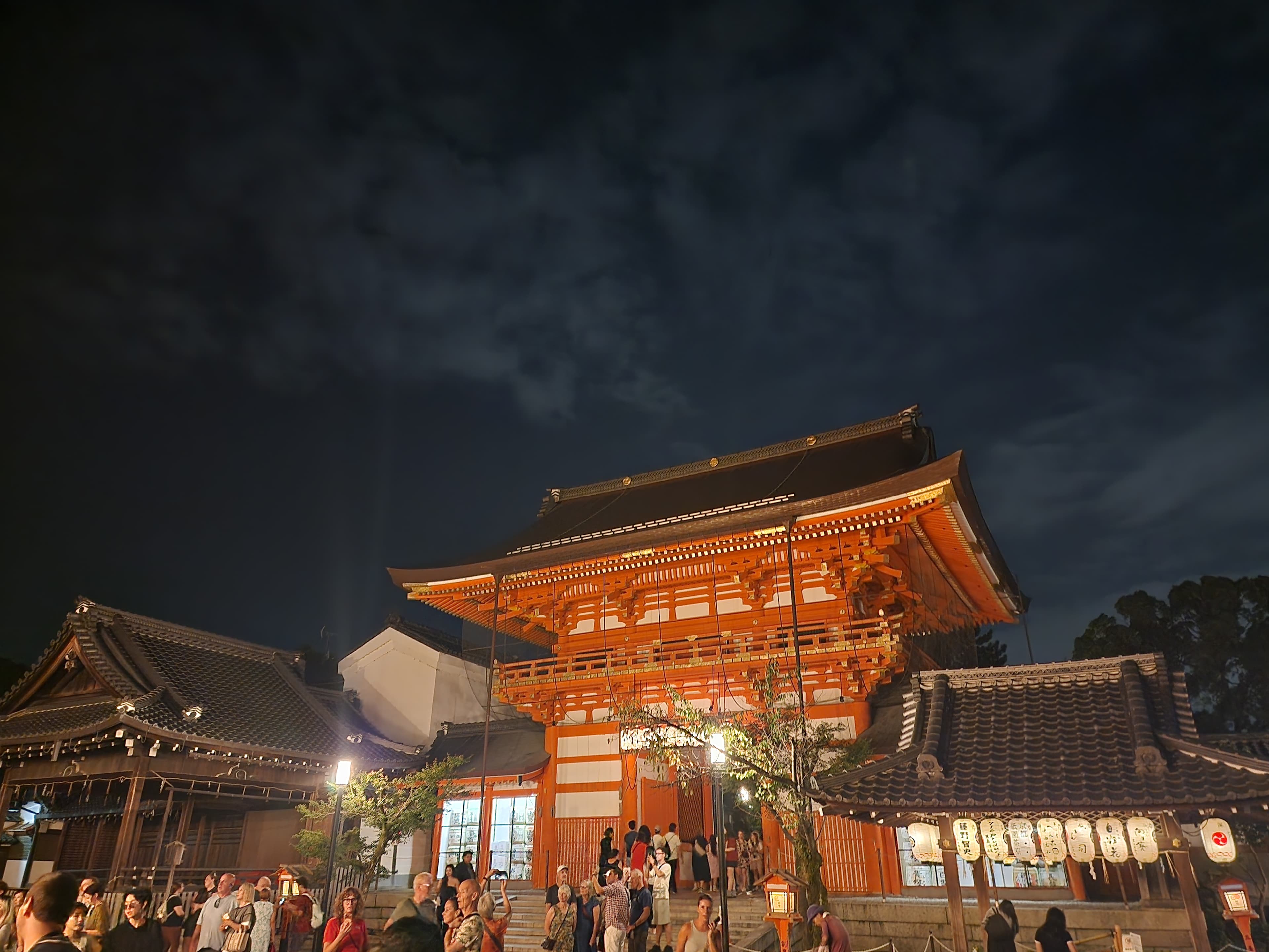 Puerta bermellón iluminada del santuario Yasaka en Kioto de noche, rodeada de gente y edificios.