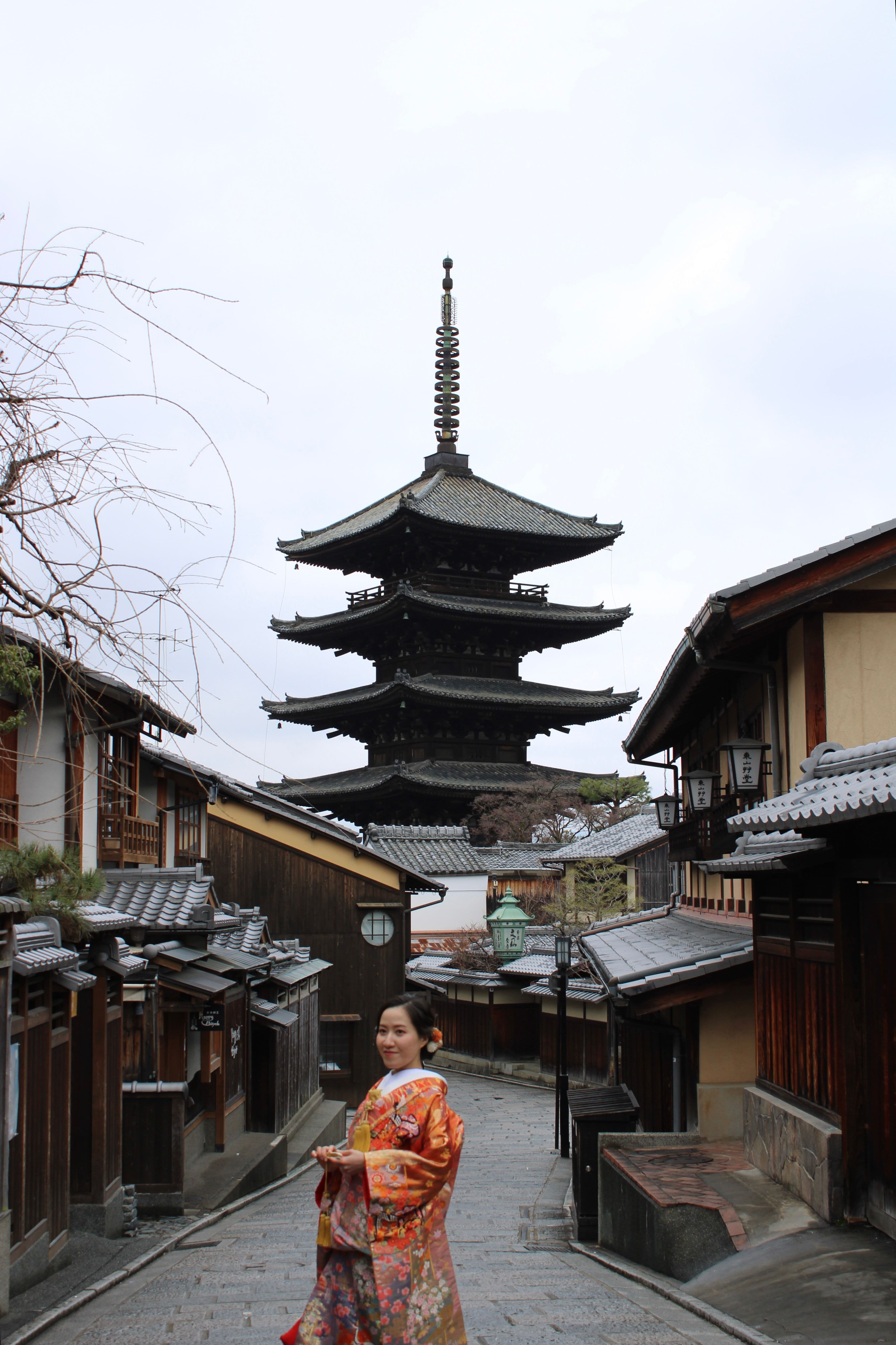 Mujer en kimono naranja caminando por calle tradicional de Kioto con pagoda histórica al fondo.
