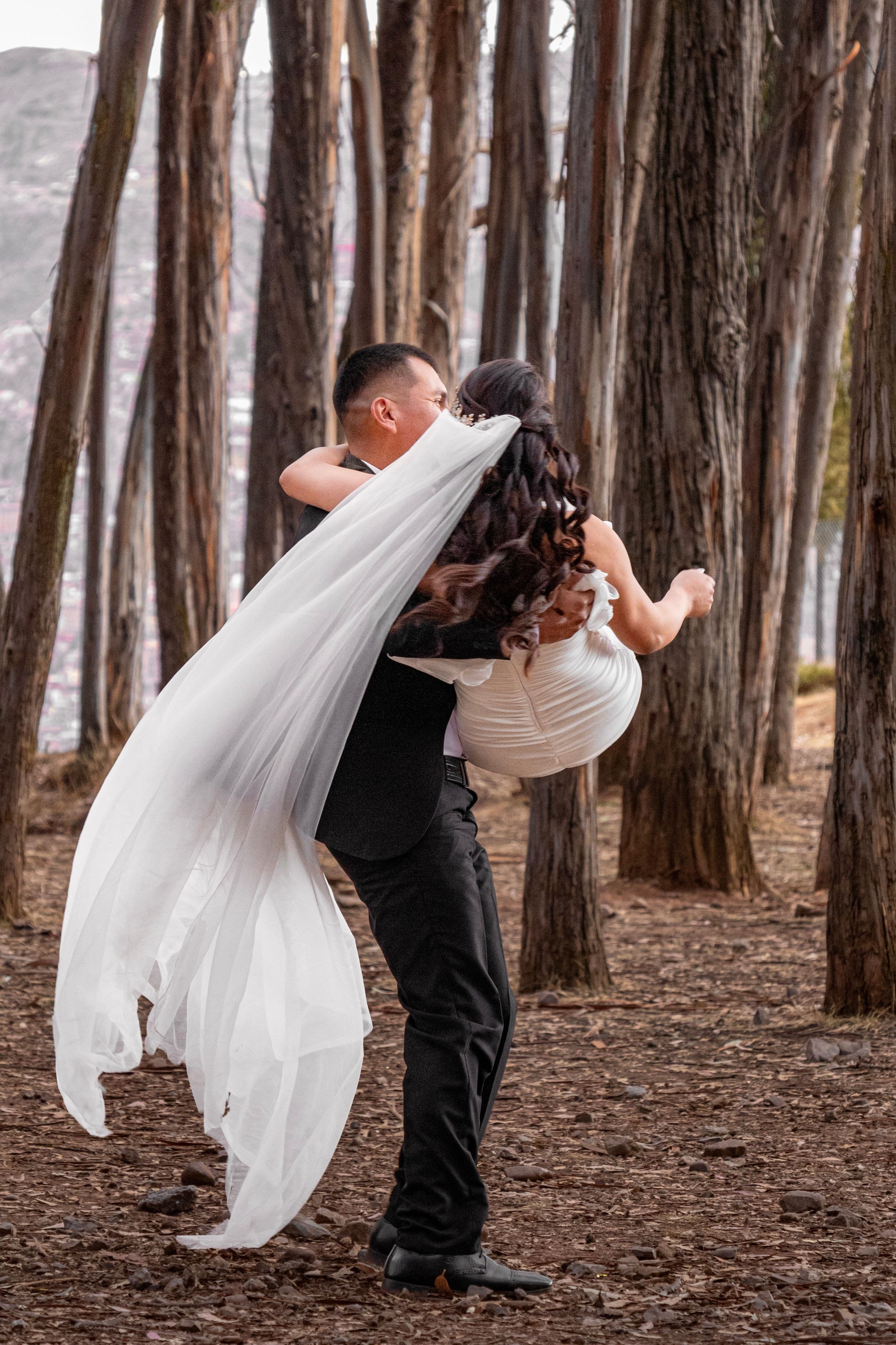 Novio cargando a su novia en un bosque con su largo velo blanco al viento.