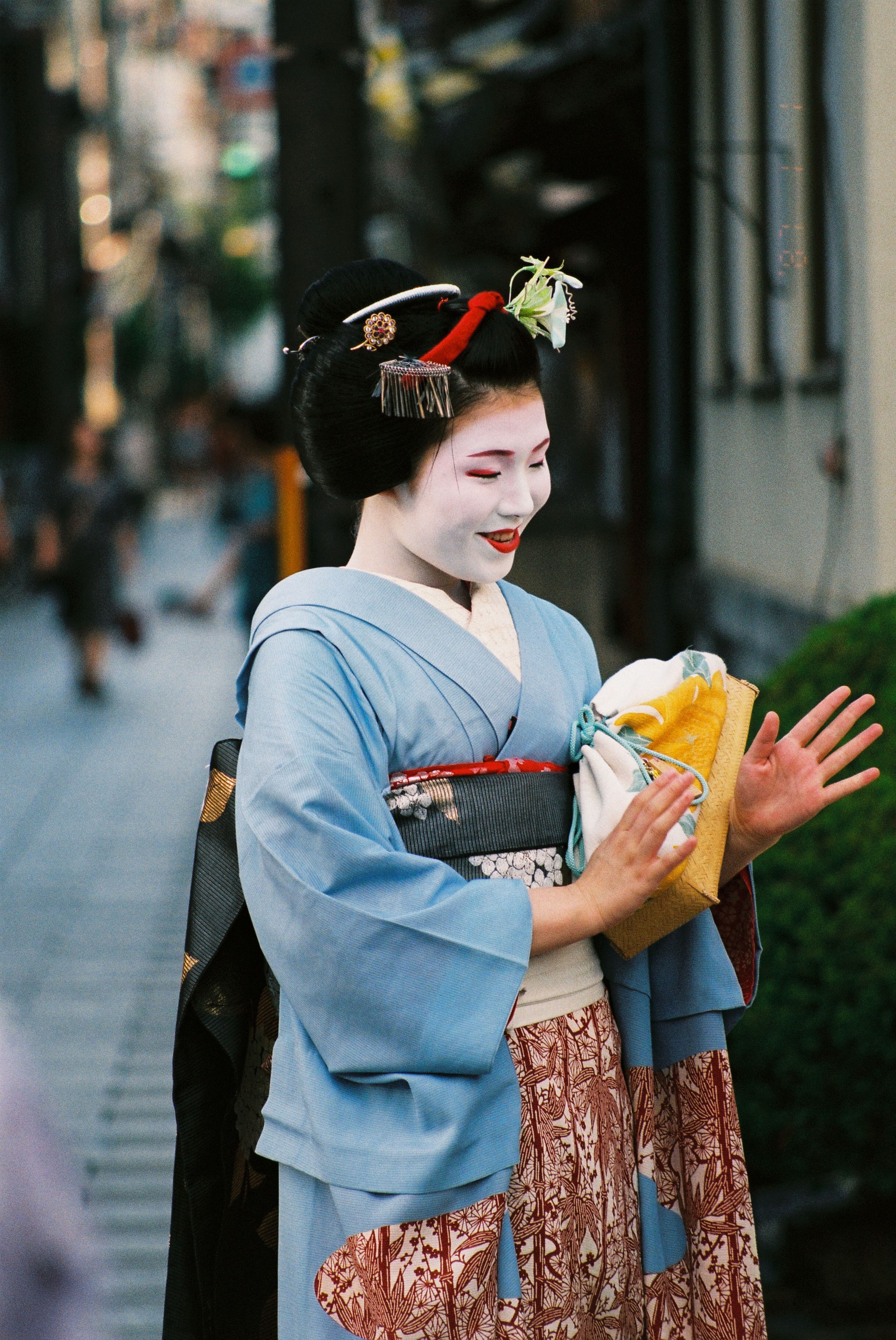 Maiko japonesa con kimono azul y maquillaje tradicional sonríe mientras sostiene un pequeño bolso.